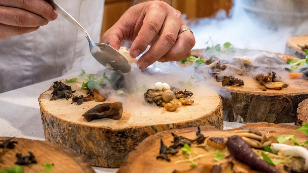 Chef plating a mushroom dish on a wood slab with dry-ice smoke in a Modern Fine Dining restaurant
